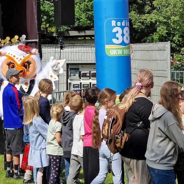 Uhlinchen steht beim Highland Gathering in Peine zwischen vielen Kindern vor der Bühne im Stadtpark. Im Hintergrund sind Pokale, schottische Dekorationen und Veranstaltungsbanner zu sehen. Die Kinder warten gespannt auf das gemeinsame Tauziehen bei den Kinder-Games.