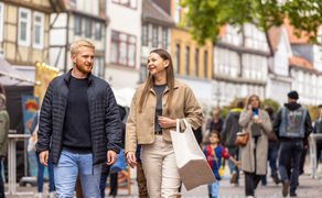 Ein Mann und eine Frau gehen lächelnd durch eine belebte Fußgängerzone mit Fachwerkhäusern, während Menschen im Hintergrund einkaufen und sich unterhalten.