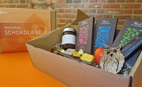 Offene Genussbox mit drei Rausch Plantagen Schokoladentafeln, einem Glas Marmelade, mehreren Pralinen und einer Holz-Eulenfigur vor Ziegelwand.
