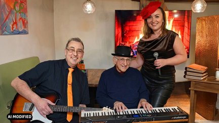Drei Personen in einem Wohnzimmer, ein Mann mit Gitarre, ein Mann am Keyboard, eine Frau mit Mikrofon.