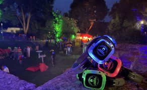 Mehrere beleuchtete Funkkopfhörer liegen auf einer Steinmauer, im Hintergrund feiern Gäste.