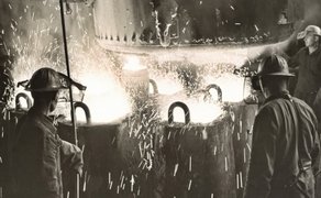 Historische Schwarz-Weiß-Aufnahme aus dem Peiner Stahlwerk. Mehrere Arbeiter stehen in einer Werkhalle beim Kokillenguss. Im Hintergrund leuchtet das flüssige, heiße Metall hell auf.