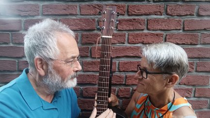 Das Musikduo Ma Y Be steht vor einer roten Backsteinwand und blickt sich lächelnd an. Zwischen ihnen halten sie gemeinsam eine Akustikgitarre. Beide tragen Brillen und haben kurzes graues Haar.