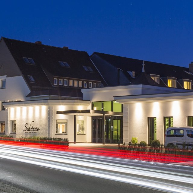 Modernes Hotelgebäude bei Nacht mit beleuchteter Fassade; vor dem Hotel ziehen Lichtspuren vorbeifahrender Autos entlang der Straße, der Himmel ist dunkelblau.