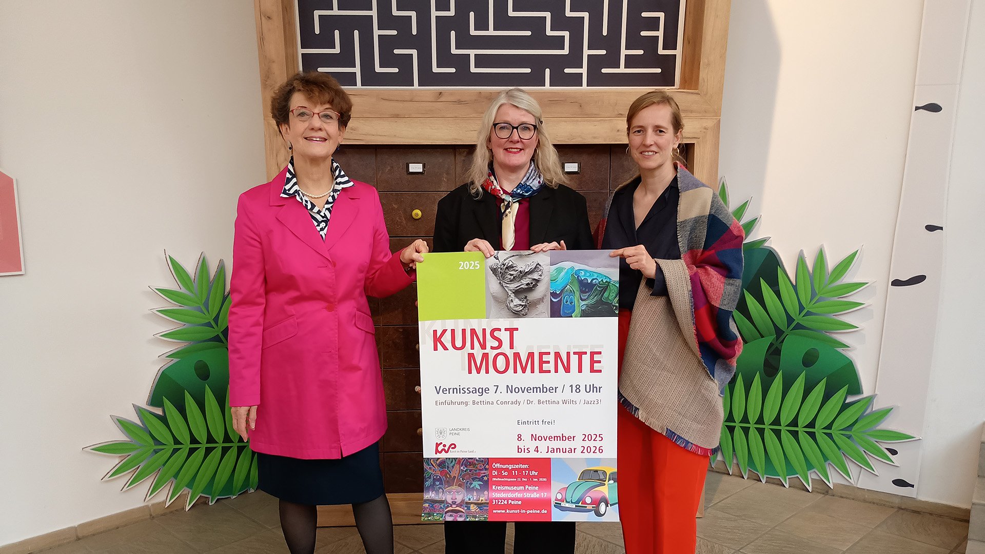 Drei Frauen stehen vor einer Holzwand mit Labyrinth-Optik und halten ein Plakat mit der Aufschrift „KUNST MOMENTE“ und „KiP“ in den Händen.