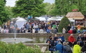 Belebtes Open-Air-Festival im Park mit vielen Besucherinnen und Besuchern rund um einen Springbrunnen, Marktständen und Buden im Hintergrund – geselliges Familienevent mit Volksfest-Atmosphäre im Grünen.