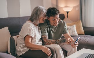 Zwei Personen sitzen gemeinsam auf einem Sofa und schauen auf einen Laptop. Eine Person zeigt auf den Bildschirm, während die andere aufmerksam zusieht.