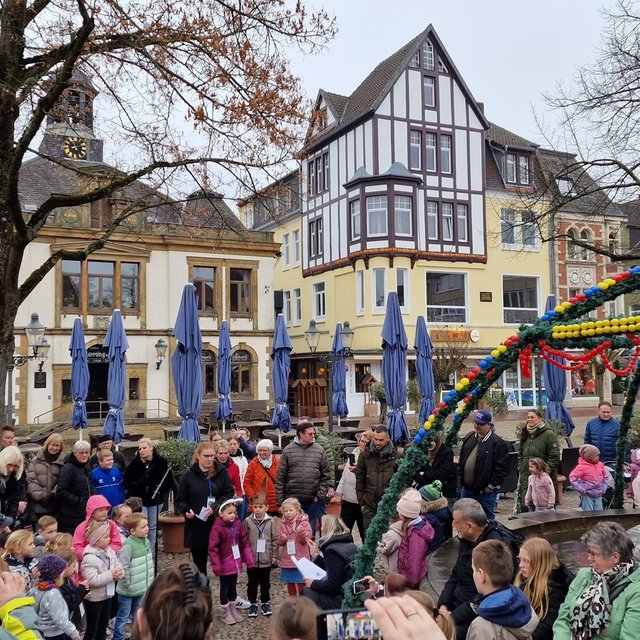 Viele Menschen, darunter zahlreiche Kinder und Erwachsene, stehen auf einem Marktplatz um einen geschmückten Brunnen herum. Der Brunnen ist mit grünen Girlanden und bunten Ostereiern dekoriert. Einige Kinder stehen im Mittelpunkt und hören aufmerksam zu, während Erwachsene zuschauen und teilweise Fotos machen. Im Hintergrund sind historische Gebäude, Bäume und geschlossene Sonnenschirme eines Cafés zu sehen.