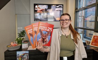 Eine Mitarbeiterin der Peine Marketing GmbH steht in der Tourist-Information und hält mehrere Exemplare des Jahresberichts 2025 in der Hand. Auf den orangefarbenen Titelseiten ist eine fliegende Eule abgebildet.