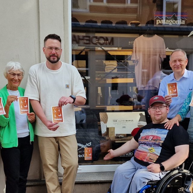 Acht Personen stehen und sitzen vor einem Geschäft in der Peiner Innenstadt und halten orangefarbene Aufkleber für mehr Barrierefreiheit in die Kamera. Eine Person sitzt im Rollstuhl, eine weitere steht mit einem Rollator vor der Gruppe. Im Schaufenster ist ein Aufkleber bereits an der Eingangstür angebracht. Die Aufnahme zeigt die Beteiligten der Initiative, die sich gemeinsam für einen barriereärmeren Zugang zu Geschäften einsetzen.