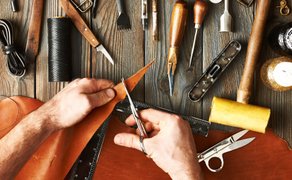 Hände schneiden mit einer Schere ein Stück Leder auf einem Holztisch mit verschiedenen Lederwerkzeugen und Garnrollen
