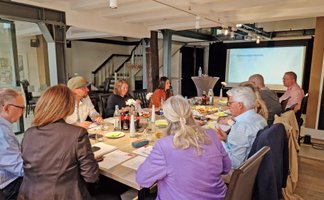 Mehrere Personen sitzen gemeinsam an einem großen Tisch in einem Veranstaltungsraum. Vor ihnen liegen Unterlagen und Notizen, auf dem Tisch stehen Getränke und kleine Snacks. Im Hintergrund ist eine Präsentationsfläche zu sehen.