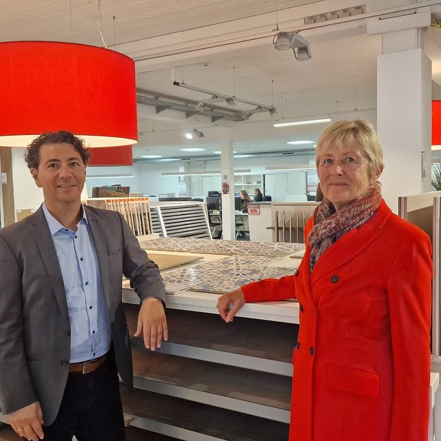 Ein Mann im Anzug und eine Frau im Mantel stehen in einem Baumarkt vor Regalen mit Fliesenmustern.