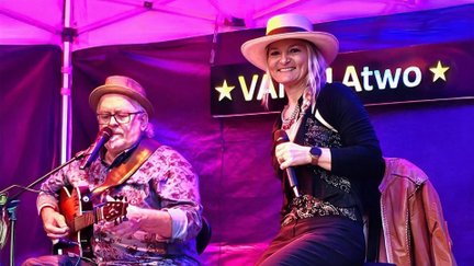 Das Duo VANILLAtwo tritt auf einer beleuchteten Bühne unter pink-violettem Licht auf. Ein Musiker sitzt mit Gitarre und singt in ein Mikrofon, während seine Partnerin mit Hut lächelnd ein Mikrofon in der Hand hält. Im Hintergrund ist ein Banner mit dem Bandnamen zu sehen.