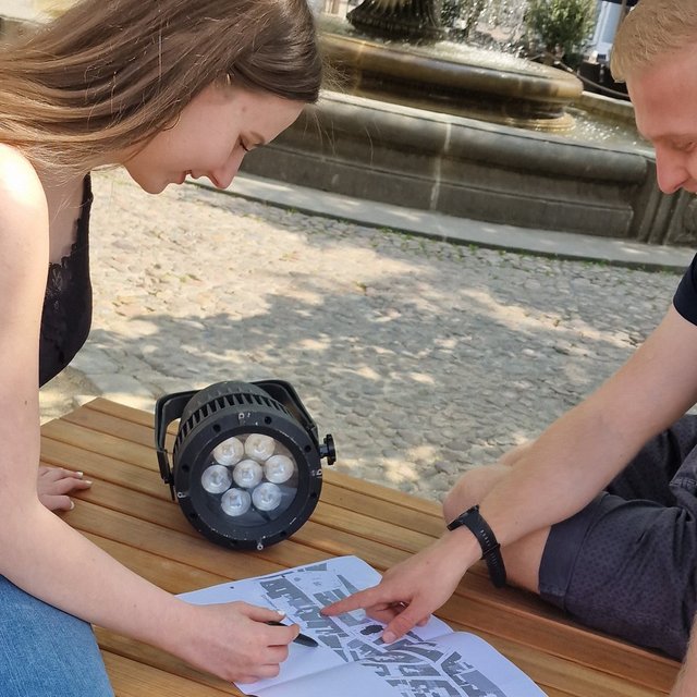 Zwei junge Erwachsene sitzen an einem Tisch im Freien, betrachten einen Lageplan und besprechen gemeinsam die Platzierung eines technischen Scheinwerfers.