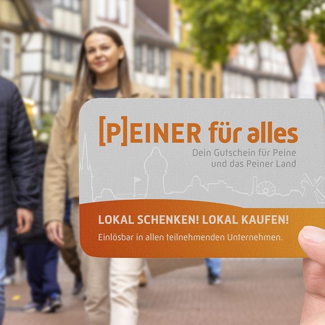 Eine Hand hält den Peiner Stadtgutschein „[P]EINER für alles“ in die Kamera. Im unscharfen Hintergrund sind Menschen in der belebten Peiner Innenstadt mit Fachwerkhäusern zu sehen.