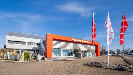 Modernes Geschäftsgebäude mit rotem Eingangsbereich und Schriftzug „Baustoff Brandes“, umgeben von bepflanzten Beeten und gepflastertem Vorplatz.