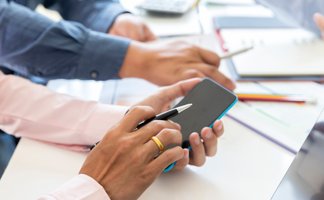 Hände von zwei Personen, eine hält ein Smartphone, die andere zeigt mit einem Stift darauf, im Hintergrund Schreibtisch mit Papieren und Laptop.