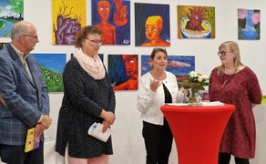 Eine Frau und ein Mann stehen vor einer Wand mit mehreren bunten Gemälden und Zeichnungen in einem Ausstellungsraum.