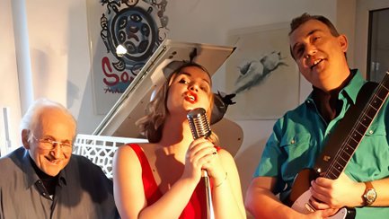 Drei Musiker der Band WestCoastJam performen gemeinsam in einem hellen Innenraum vor einem weißen Flügel. Eine Sängerin im roten Kleid singt in ein Retro-Mikrofon, während sie von einem Pianisten und einem Gitarristen begleitet wird. Warmes Licht sorgt für eine stimmungsvolle Jazz-Atmosphäre.