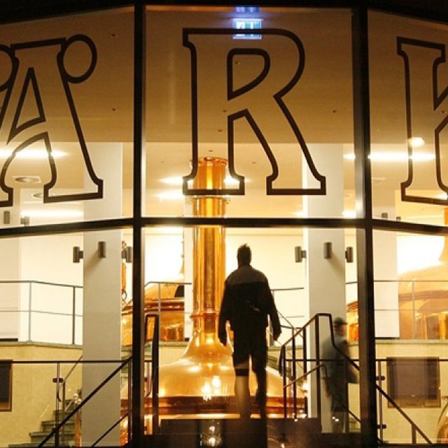 Blick durch die Glasfassade der Härke Braumanufaktur Peine auf die beleuchteten Kupferkessel im Inneren der Brauerei. Eine Person betritt den zentralen Brauraum und unterstreicht die moderne Verbindung von Braukunst, Architektur und Biergenuss aus Peine.
