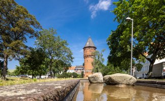 Im Vordergrund verläuft ein flacher Wasserlauf mit Steinen, dahinter erhebt sich ein runder Backsteinturm mit spitzem Dach. Der Turm ist von Bäumen und Gebäuden umgeben. Die Szene ist bei sonnigem Wetter aufgenommen, der Himmel ist blau. Die Perspektive ist niedrig und lenkt den Blick entlang des Wassers auf den historischen Turm.