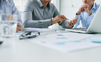 Mehrere Personen sitzen an einem Tisch in einer Besprechung, diskutieren Unterlagen und arbeiten gemeinsam mit Laptop und Notizen in einer professionellen Büroumgebung.