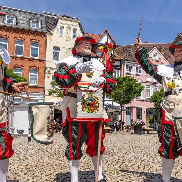 Drei Männer in historischen, grün-roten Uniformen mit Trommeln stehen auf einem gepflasterten Marktplatz in Peine und spielen fröhlich Musik vor bunten Altstadthäusern bei sonnigem Wetter.