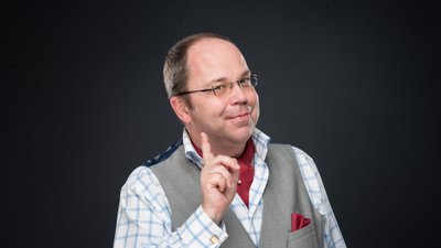 Mann mit grauem Weste, kariertem Hemd und rotem Einstecktuch hebt den Zeigefinger und trägt ein rotes Halstuch.