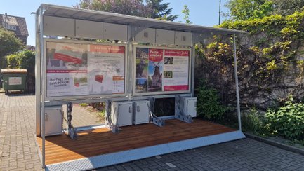 Überdachte Bikeport-Anlage in der Peiner Innenstadt mit Infotafeln, Fahrradboxen und Lademöglichkeiten, daneben E-Ladesäulen und historische Backsteingebäude im Hintergrund.