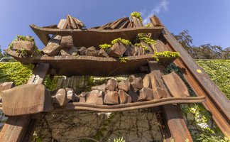Rostige Stahlskulptur mit aufgeschichteten Metall- und Steinformen, überwachsen von Pflanzen, im öffentlichen Raum in Peine.