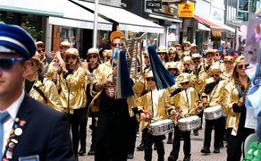 Musiker in goldfarbenen Jacken mit Trommeln und Blasinstrumenten ziehen in einer belebten Einkaufsstraße an Zuschauern vorbei.