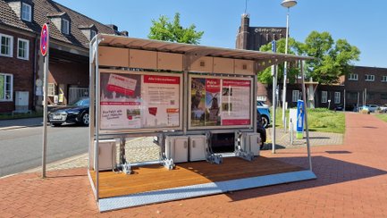 Überdachte Bikeport-Anlage in der Peiner Innenstadt mit Infotafeln, Fahrradboxen und Lademöglichkeiten, daneben E-Ladesäulen und historische Backsteingebäude im Hintergrund.