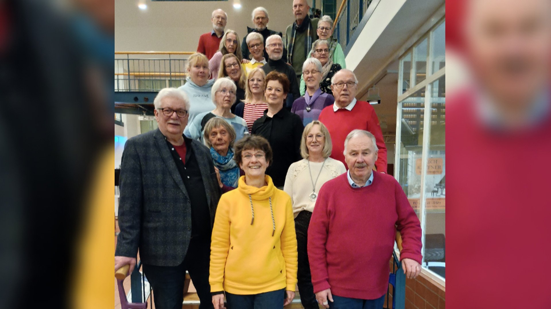 Gruppe von Menschen unterschiedlichen Alters in einem Innenraum mit Treppe und Geländer im Hintergrund.