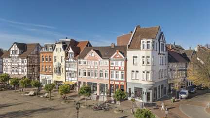 Blick auf den Historischen Marktplatz in Peine mit einer Reihe farbenfroher, teils denkmalgeschützter Fachwerk- und Altstadthäuser unter blauem Himmel. Der gepflasterte Platz ist von kleinen Bäumen, Sitzgelegenheiten und Fahrrädern geprägt.
