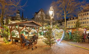Weihnachtlich beleuchteter Marktplatz in Peine mit geschmückten Tannenbäumen, Holzbank mit Schild 'Kaufmannsgilde zu Peine' und Fachwerkhäusern im Hintergrund.