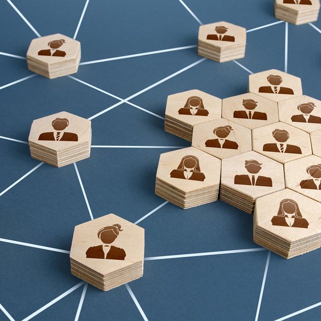 Holzplättchen mit stilisierten Personen in Anzügen, verbunden durch weiße Linien auf blauem Hintergrund, symbolisieren Netzwerk.