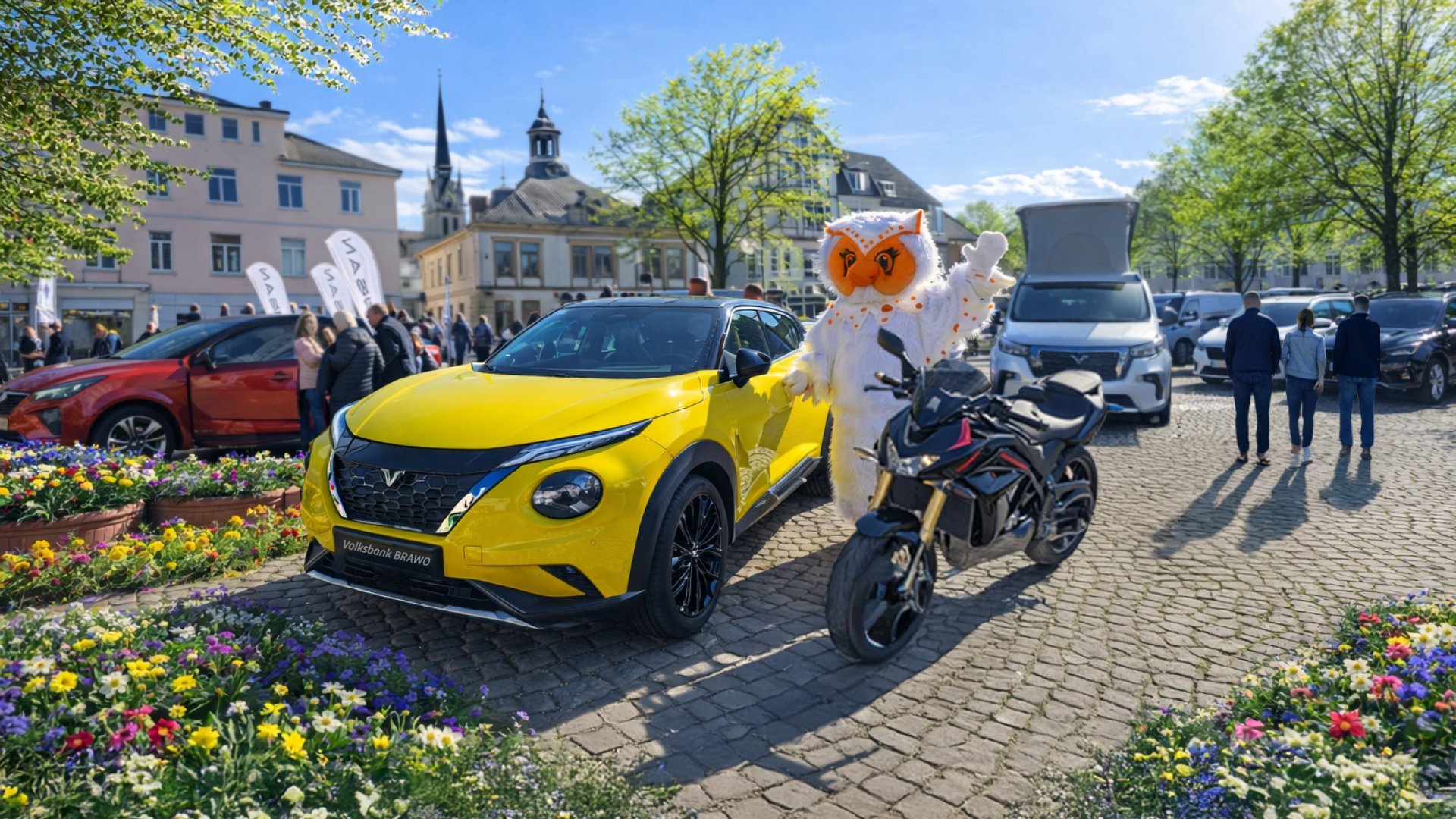 Gelbes Auto und schwarzes Motorrad auf gepflastertem Platz mit Person im Eulen-Kostüm und mehreren Besuchern bei sonnigem Wetter.