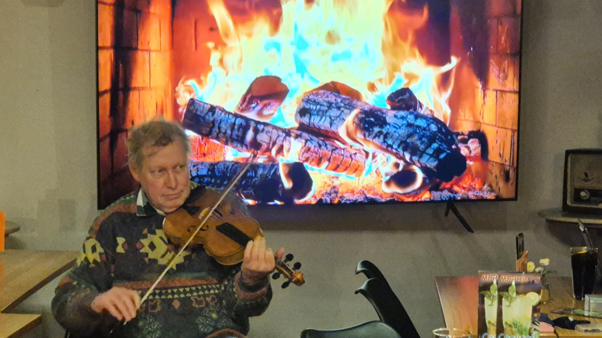 Das Bild zeigt einen Musiker, der Geige spielt. Im Hintergrund wird ein Lagerfeuer auf einem Fernsehgerät gezeigt. 