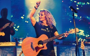 Eine Frau steht auf einer Bühne, hält eine Gitarre in der Hand und hebt den rechten Arm in die Höhe.