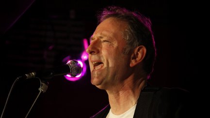 Rockmusiker Helge Holm singt auf einer Bühne leidenschaftlich in ein Mikrofon. Im Hintergrund sorgen pinkfarbene Bühnenlichter und dunkle Konzertatmosphäre für eine stimmungsvolle Beleuchtung seines Auftritts.