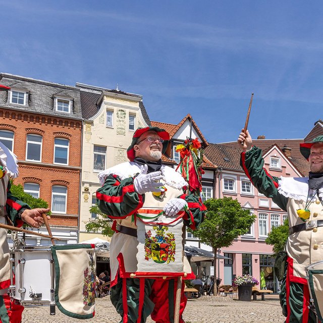 : Drei Männer in historischen Uniformen des Peiner Freischießens spielen auf Trommeln auf einem Marktplatz.