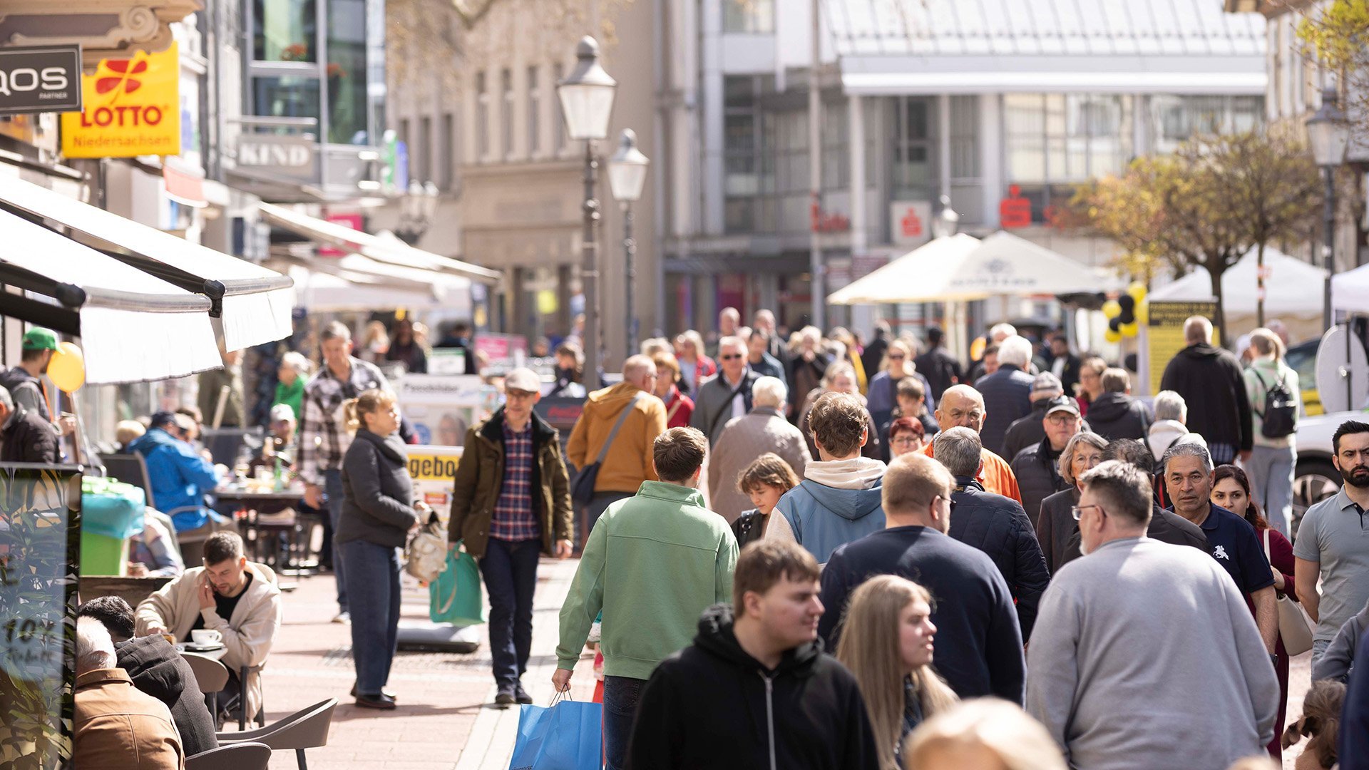 Belebte Fußgängerzone in einer Innenstadt mit zahlreichen Menschen beim Einkaufen und Flanieren, Außengastronomie, Geschäften und Sonnenschirmen bei sonnigem Wetter.