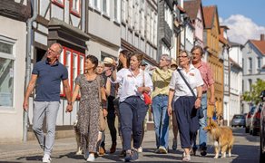 Gruppe von Menschen bei einer Stadtführung in einer historischen Altstadtstraße, die aufmerksam den Erklärungen der Guide folgen; Fachwerkhäuser, enge Gasse und ein Hund begleiten die Szene bei sonnigem Wetter.