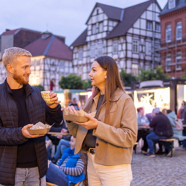 Ein Mann und eine Frau stehen mit Getränken auf einem belebten Platz, im Hintergrund sitzen und stehen weitere Menschen vor Fachwerkhäusern.