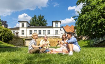 Vier Personen sitzen auf einer Picknickdecke auf einer Rasenfläche und stoßen mit Getränken an. Im Hintergrund stehen ein historisches Gebäude, eine Natursteinmauer und große Bäume unter blauem Himmel.