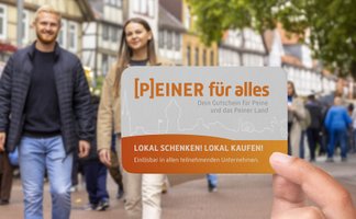 Eine Hand hält den Peiner Stadtgutschein „[P]EINER für alles“ in die Kamera. Im unscharfen Hintergrund sind Menschen in der belebten Peiner Innenstadt mit Fachwerkhäusern zu sehen.