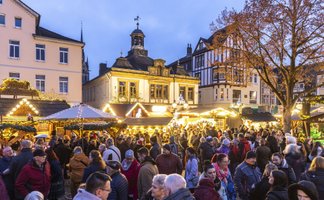 Belebter Weihnachtsmarkt mit festlich beleuchteten Ständen vor historischen Fachwerkhäusern und einem barocken Gebäude im Abendlicht.