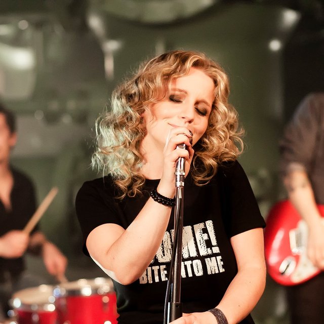 Sängerin mit lockigem Haar hält Mikrofon, im Hintergrund Schlagzeuger und Gitarrist mit roter E-Gitarre auf Bühne.