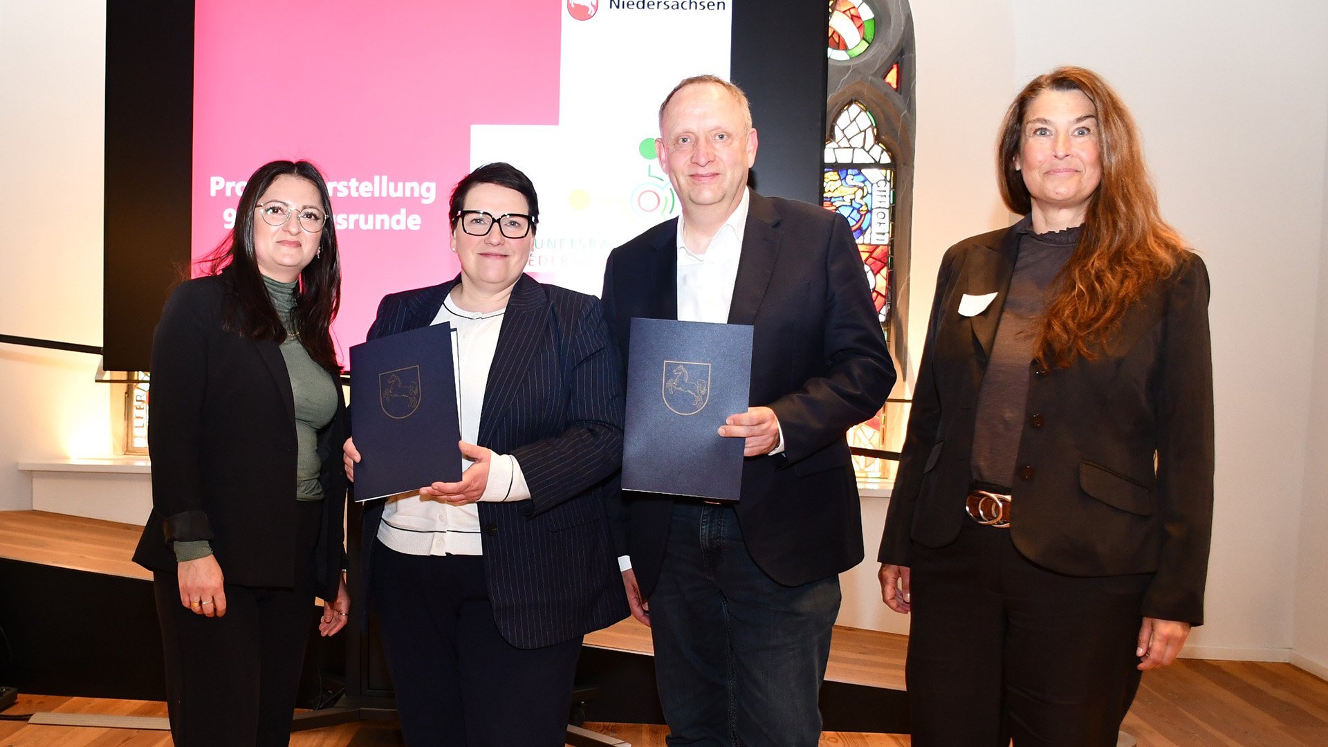 Vier Personen stehen bei der Übergabe von Förderbescheiden des Programms „Zukunftsräume Niedersachsen“ vor einem Bildschirm mit Präsentationsfolie in einem historischen Saal.